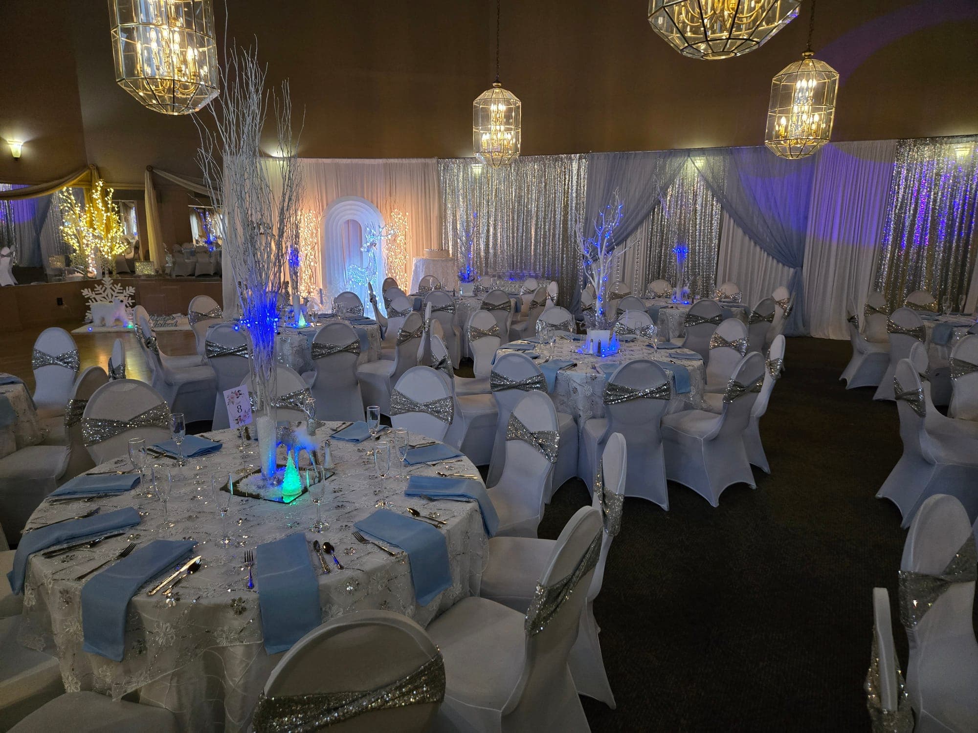 Winter wonderland party with white chair covers, silver sequin sashes, blue napkins and lit white branch centerpieces in Delran NJ