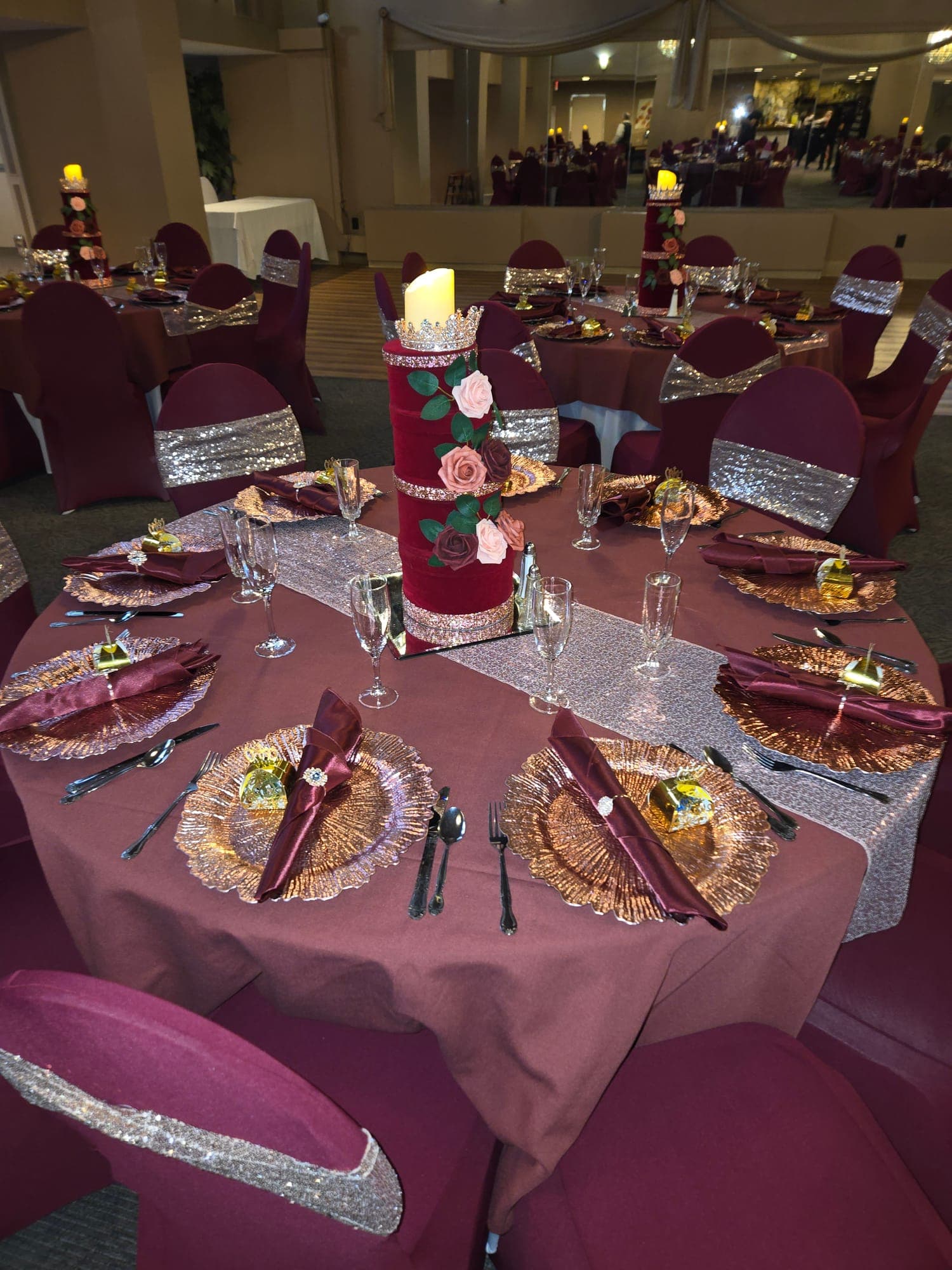Round table in burgundy linens with gold chargers, silver sequin runner and a rose-wrapped pillar candle centerpiece