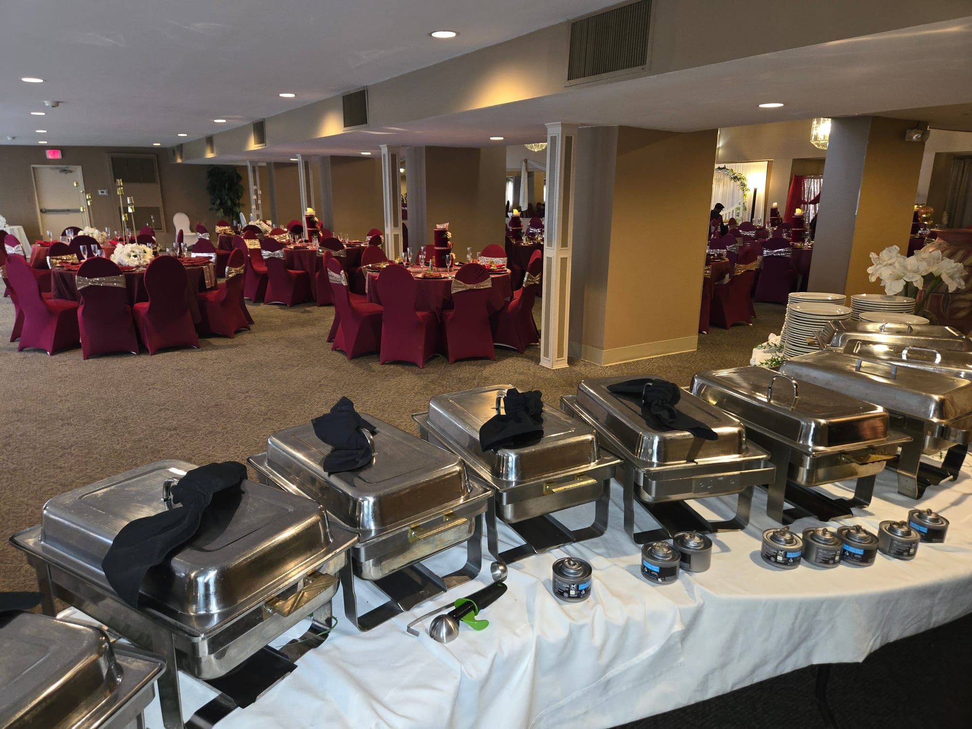 Line of polished chafing dishes ready for a buffet in a banquet hall set with burgundy chair covers