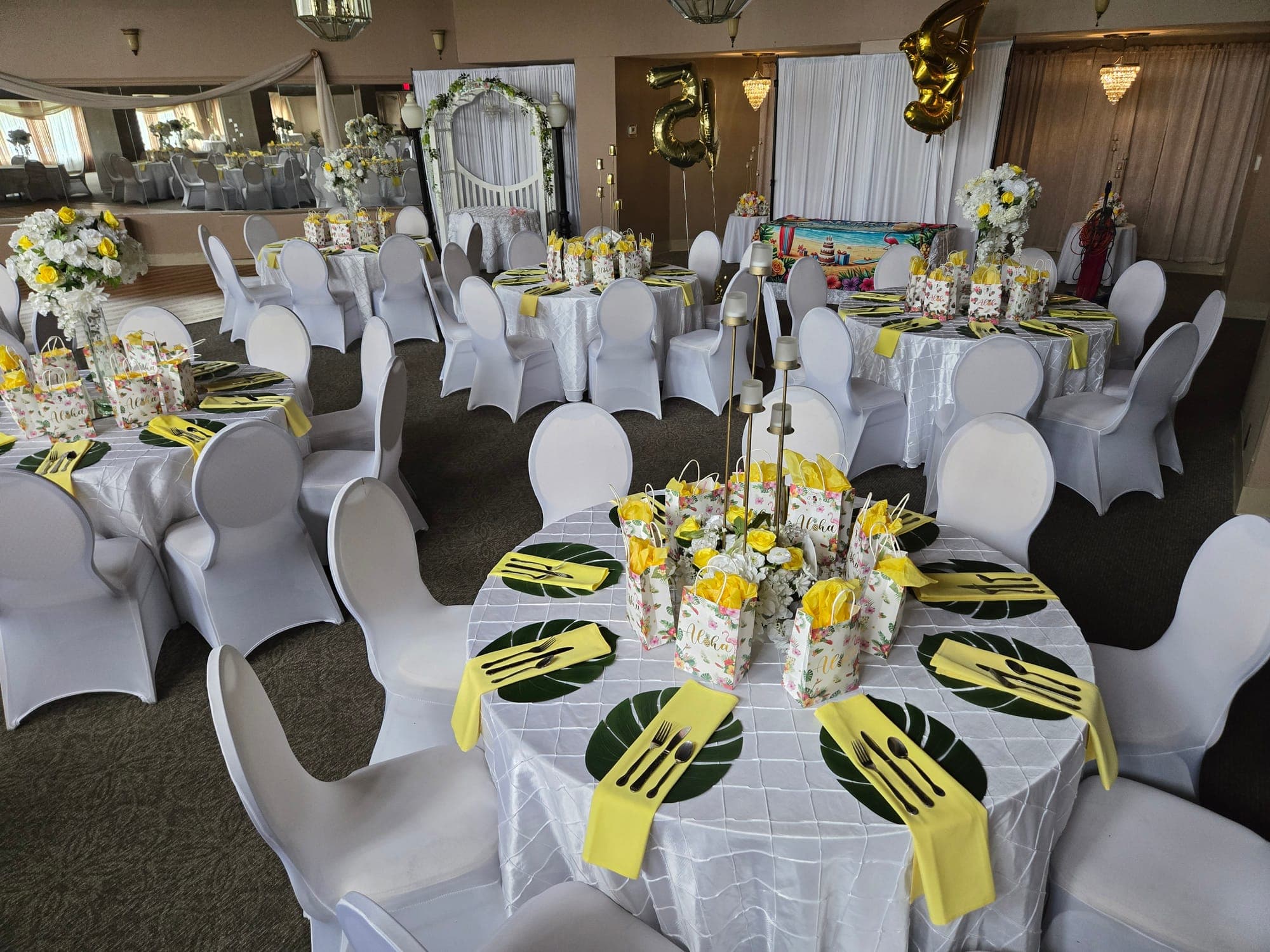 Luau-themed party room with palm-leaf placemats, yellow napkins, aloha favor bags and gold number balloons