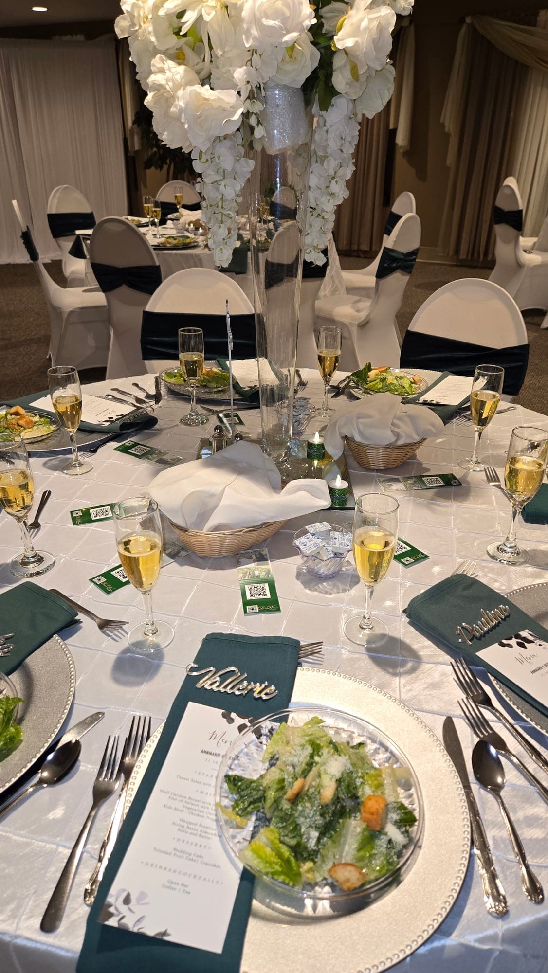 Place setting with Caesar salad, champagne flutes, a printed menu and emerald napkins beneath a tall white floral centerpiece