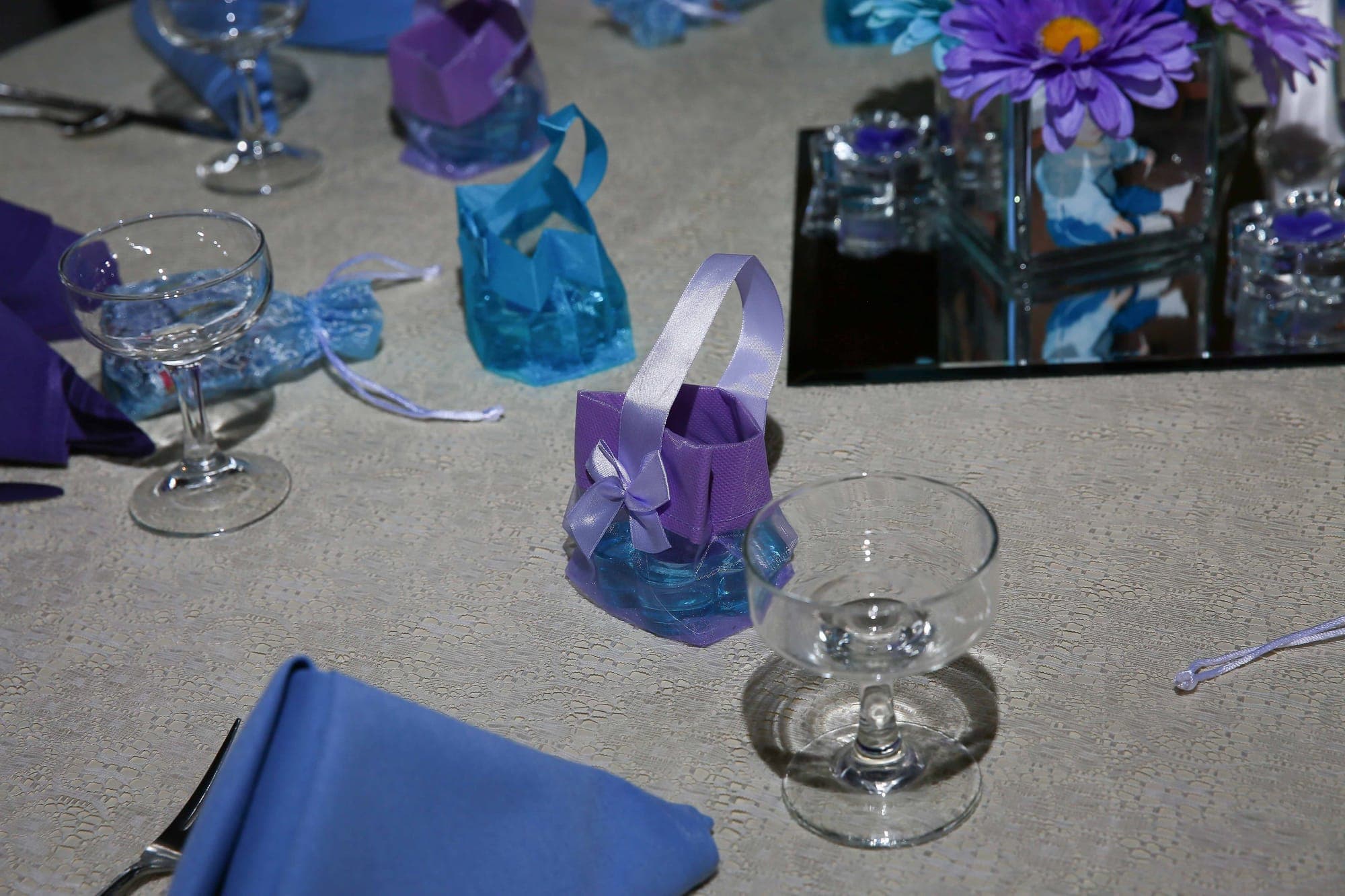 Lavender and turquoise favor baskets beside coupe glasses and blue napkins on a lace tablecloth